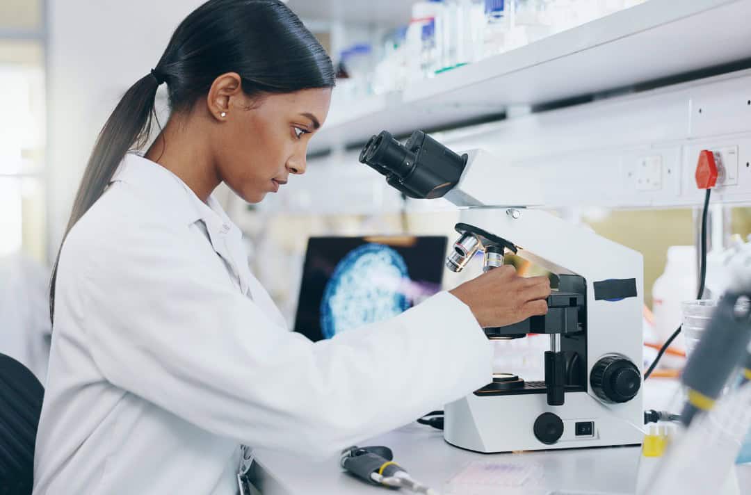 A woman in a while lab coat looking through a microscope in a laboratory setting.