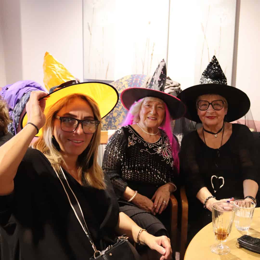 Three women sat at a table with witches hats on, celebrating Halloween.