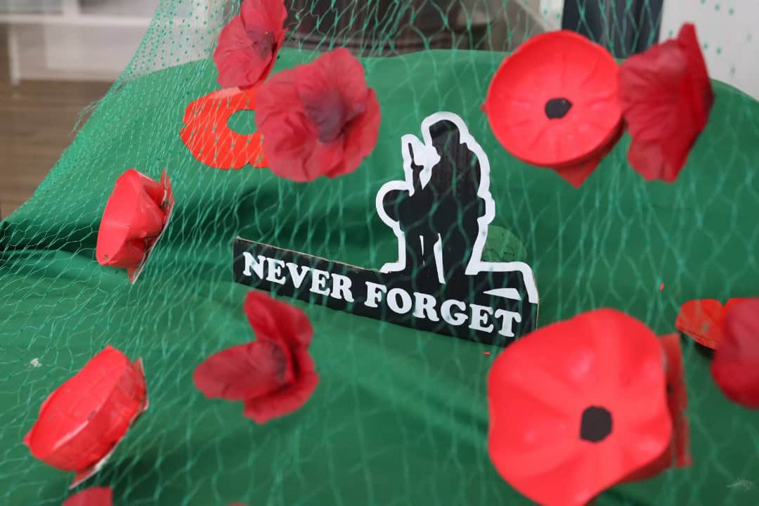 Silhouette of a soldier kneeling on one knee with the text 'Never Forget' underneath it. This is stood on a green table with poppies surrounding it.