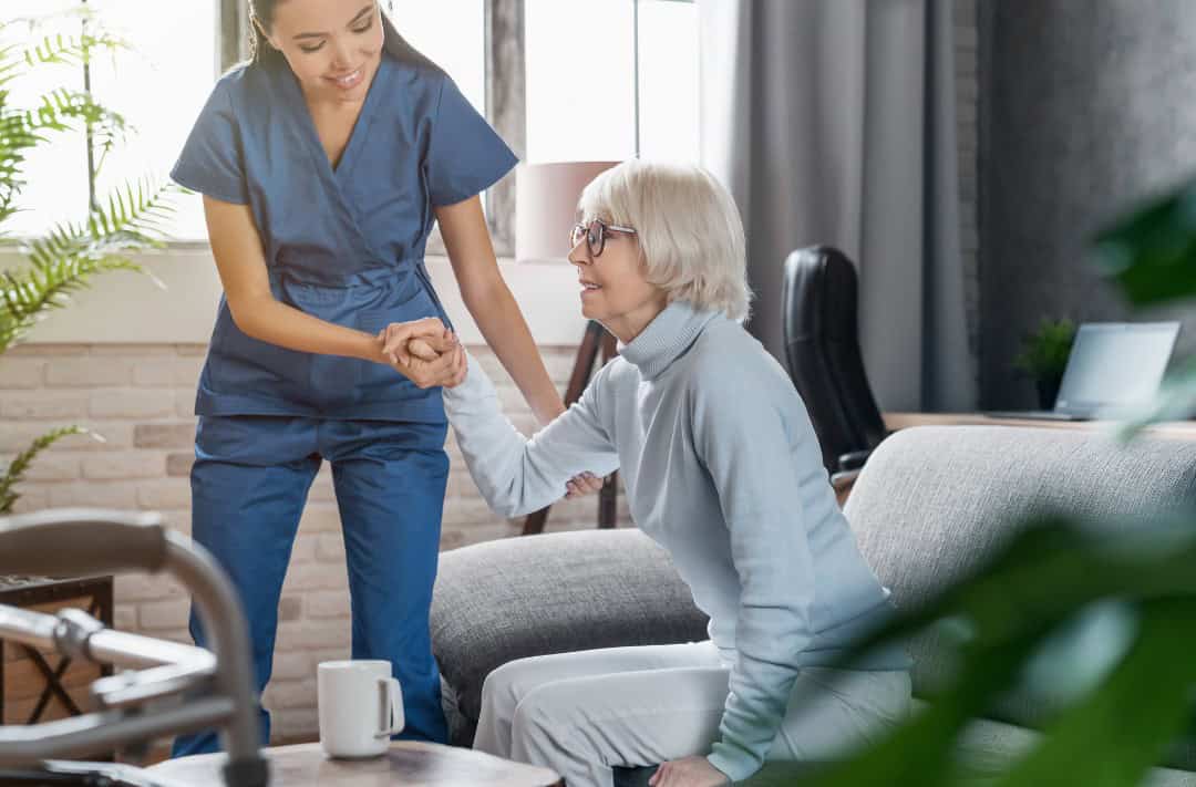 A healthcare worker in blue scrubs assisting an elderly patient who is sitting on the edge of a sofa in a well-lit living room.